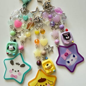 May include: Three star-shaped keychains featuring Sanrio characters. Each keychain includes a metal clasp, colorful beads in shapes of hearts, stars, and fish, and a small handheld game console charm alongside a large star-shaped character pendant.