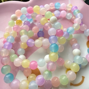 May include: A collection of pastel-coloured beaded bracelets displayed on a pink dish. The bracelets feature a variety of soft colours, including pink, yellow, blue, green, and purple. The beads are round and appear to be made of a translucent material.