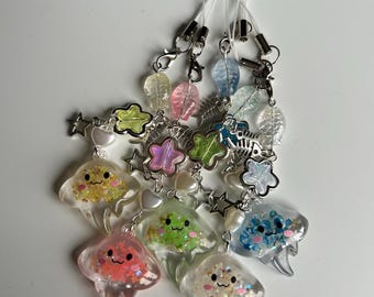 May include: A set of seven colorful, glittery, resin phone charms shaped like clouds with cute faces. Each charm has a silver star, a pearl bead, and a silver charm attached to it. The charms are attached to white phone straps.