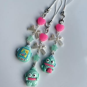 May include: Three novelty phone charms featuring light blue monster figures, pink heart beads, white bows, and star-shaped accents. One charm has the word "Hello!" on it. Each charm has a silver clasp and white cord.