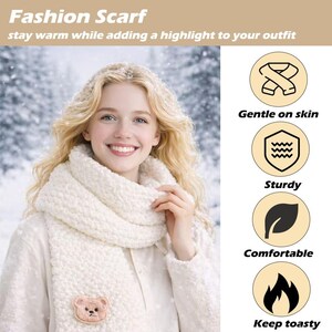 May include: A thick, cream-colored chunky knit scarf featuring a small, embroidered brown bear face patch at the bottom. The scarf is worn wrapped around the neck, paired with a matching cream-colored textured top against a snowy background.
