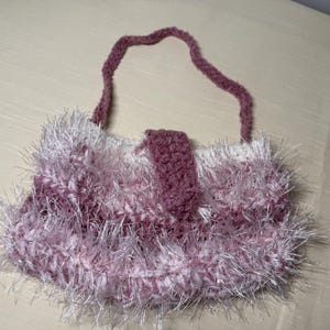 May include: A handmade, fluffy crochet bag in shades of pink and white. The bag features a textured, fringed design and a matching handle. A flap with a button closure secures the bag's contents. The bag is a unique accessory.