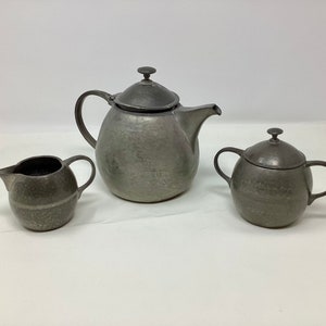 Peut inclure: Un service à thé en argent composé d'une théière, d'un pot à crème et d'un sucrier. L'ensemble a une finition martelée et un design classique.