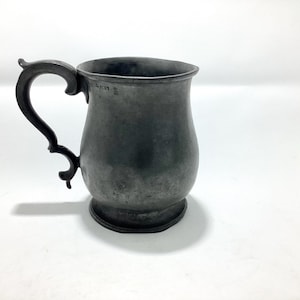 May include: A pewter tankard with a decorative handle. The mug has a bulbous body and a flared rim. The handle is ornate with a scroll design. The tankard is a dark grey colour and appears to be antique.
