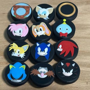 May include: A collection of twelve black circular thumbstick covers for gaming controllers. Each cover features a raised, colorful silicone character or icon from the Sonic the Hedgehog series, including Sonic, Tails, Knuckles, Amy, Shadow, and others.