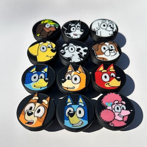 May include: A collection of twelve black circular accessories, each featuring a different cartoon dog face. The dogs are depicted in various colors, including yellow, blue, red, and brown, with distinct facial expressions and designs. The accessories are arranged in a grid.