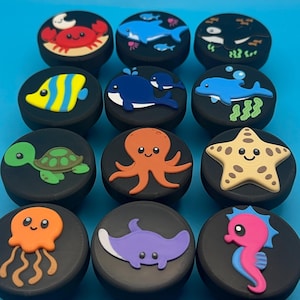 May include: A collection of black, round decorative knobs featuring various sea creatures. Designs include a red crab, blue whale, yellow fish, green turtle, orange octopus, starfish, jellyfish, stingray, and seahorse. Each knob has a different sea creature design.