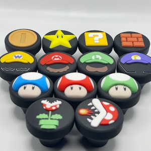May include: A selection of black cabinet knobs with vibrant, cartoon designs. The knobs showcase familiar imagery including a gold coin, a yellow star, a question mark, and various mushroom and character designs. These knobs are likely for furniture or decorative use.