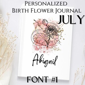 May include: A personalised journal with a pink and gold watercolour design featuring a lotus flower. The journal is titled "July" and has the name "Abigail" written in a cursive font. The text "FONT #1" is also visible at the bottom of the journal.