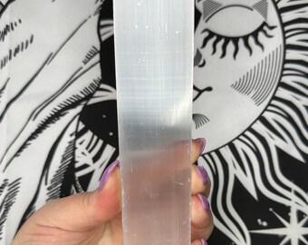 May include: A rectangular, translucent white selenite crystal wand. The crystal is held against a black and white tapestry with a sun and star design. The wand is approximately 1 inch wide and 6 inches long.