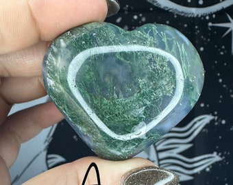 May include: Green and white heart-shaped agate stone with a white circle pattern. The stone is being held in a hand.