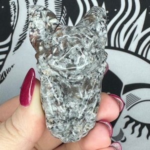 May include: A hand holding a small, gray, carved stone bat figurine. The bat has a detailed, textured surface and is made of granite.