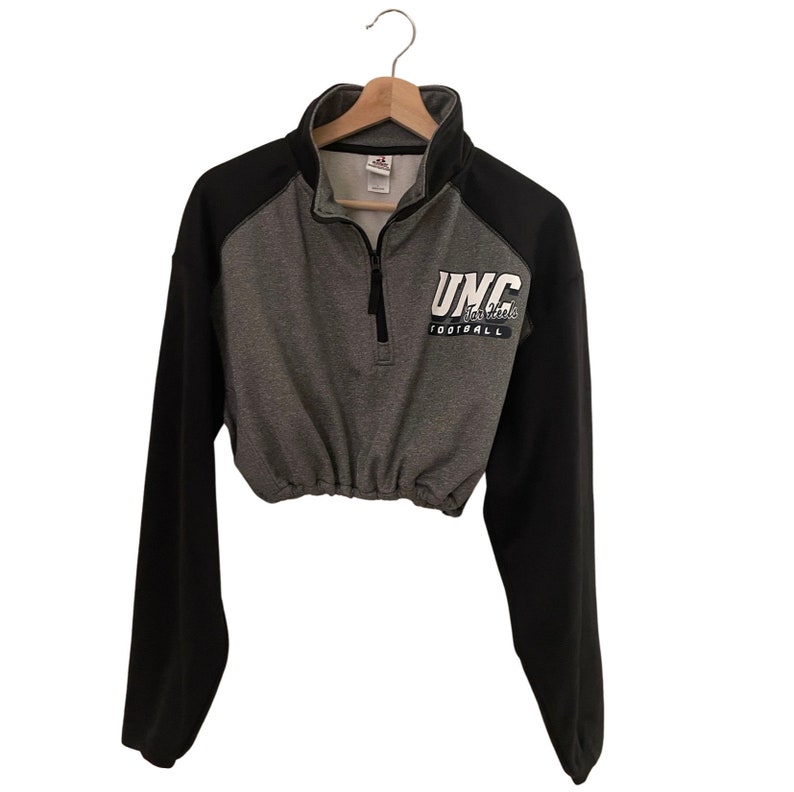 Op de afbeelding: Een grijs en zwart getailleerd sweatshirt met een wit UNC Tar Heels Football-logo op de voorkant.