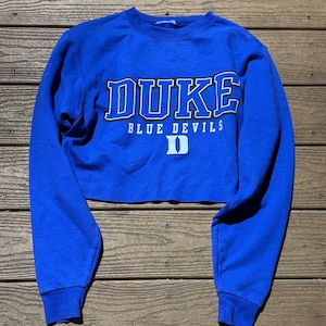 May include: A royal blue long-sleeve cropped sweatshirt featuring the text DUKE in white with a black outline, the words BLUE DEVILS in white, and a white letter D logo centered on the front. The garment has a crew neckline and ribbed cuffs.