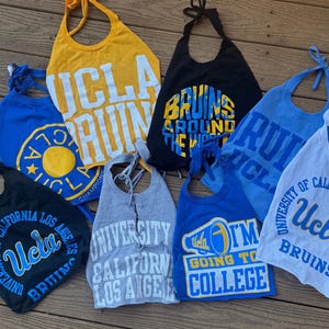 May include: Assorted UCLA Bruins bibs in various colors, including yellow, blue, black, and gray. Each bib features UCLA graphics and text, with tie closures at the neck.