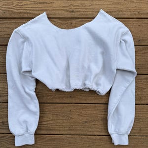 May include: A white long-sleeve cropped sweatshirt featuring a wide, open neckline and a gathered, elasticized hemline. The garment is constructed from a soft, fleece-like material with ribbed cuffs at the wrists and a clean, solid white finish.