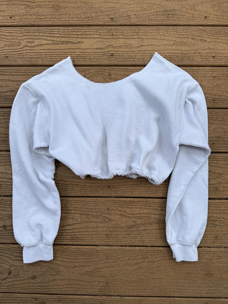 May include: A white long-sleeve cropped sweatshirt featuring a wide, open neckline and a gathered, elasticized hemline. The garment is constructed from a soft, fleece-like material with ribbed cuffs at the wrists and a clean, solid white finish.