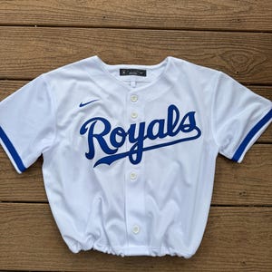 May include: A white short-sleeved button-down baseball jersey featuring the word Royals in blue script across the chest. The garment includes a small blue Nike swoosh logo on the right chest, blue trim on the sleeves, and a gathered elastic hem.