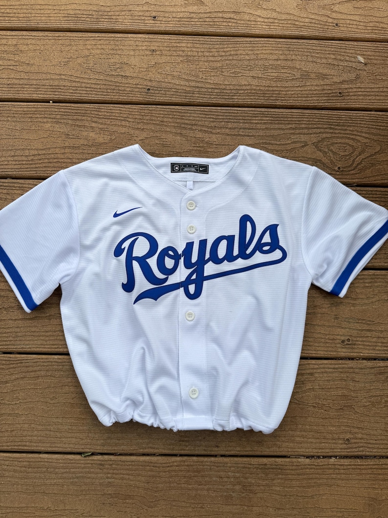 May include: A white short-sleeved button-down baseball jersey featuring the word Royals in blue script across the chest. The garment includes a small blue Nike swoosh logo on the right chest, blue trim on the sleeves, and a gathered elastic hem.