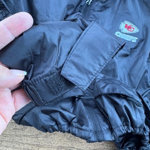 May include: Black nylon jacket with a zipper closure and a drawstring hem. The jacket features a Chiefs logo on the sleeve. The sleeve has a velcro closure. The jacket is made of a lightweight material.