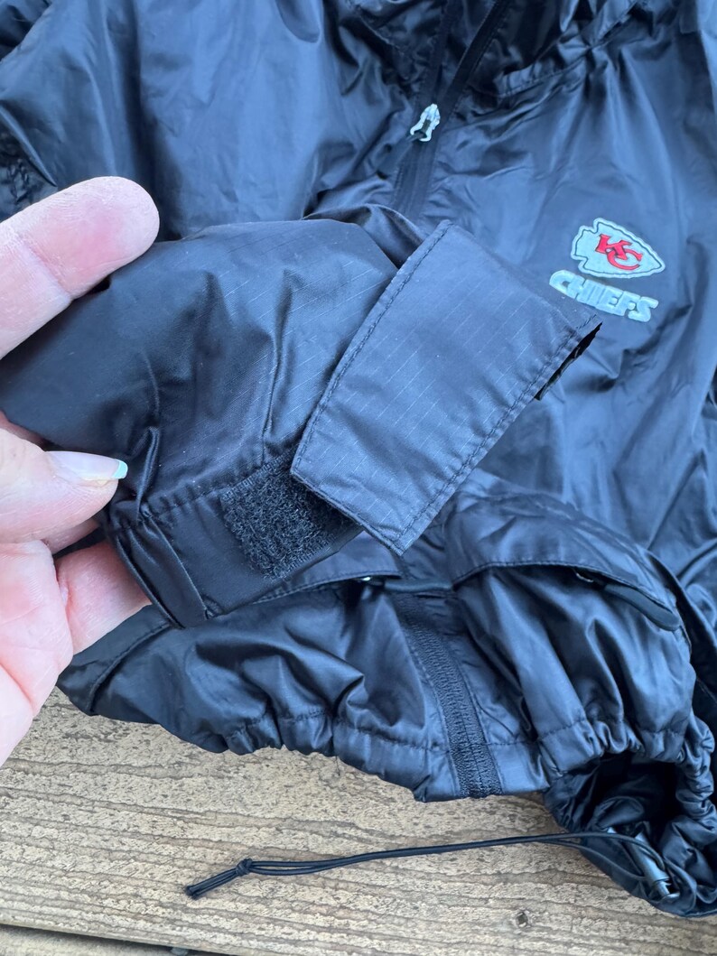 May include: Black nylon jacket with a zipper closure and a drawstring hem. The jacket features a Chiefs logo on the sleeve. The sleeve has a velcro closure. The jacket is made of a lightweight material.