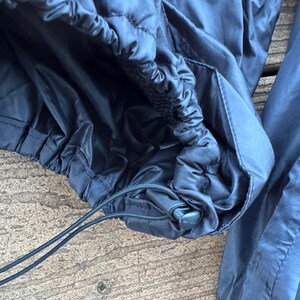 May include: Close-up of black nylon pants with an elastic waistband and drawstring. The pants have a quilted texture and a shiny finish. The drawstring is black and the pants appear to be new.