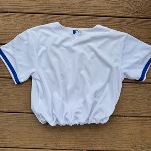 May include: A white short-sleeved jersey with blue trim on the sleeves and an elasticized hem. The back of the neckline features a small, rectangular Major League Baseball logo patch with a blue, white, and red silhouette of a batter.