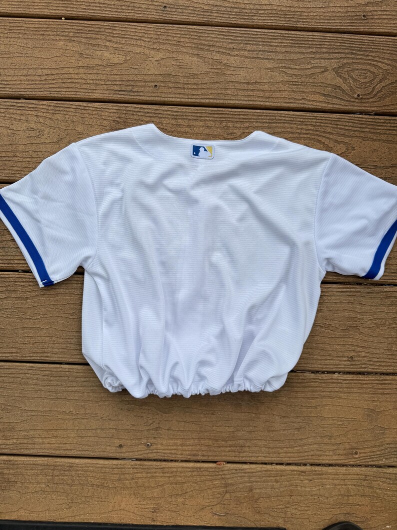 May include: A white short-sleeved jersey with blue trim on the sleeves and an elasticized hem. The back of the neckline features a small, rectangular Major League Baseball logo patch with a blue, white, and red silhouette of a batter.