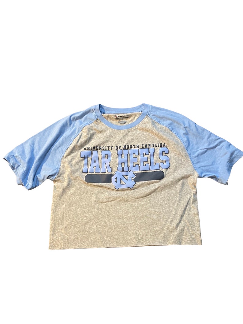 May include: A light beige and blue short-sleeved t-shirt with the text "UNIVERSITY OF NORTH CAROLINA TAR HEELS" in blue. The shirt has blue sleeves and a cropped hemline. The University of North Carolina logo is also visible.