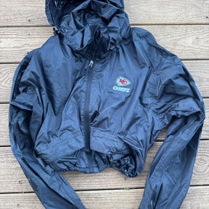 May include: Black cropped windbreaker jacket with a hood and a zipper closure. The jacket features the Kansas City Chiefs logo on the chest. The jacket has long sleeves and a drawstring at the waist.