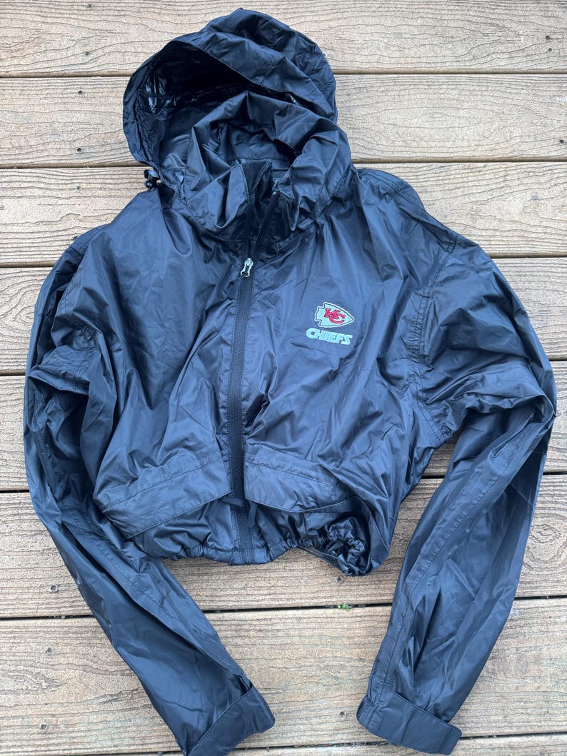 May include: Black cropped windbreaker jacket with a hood and a zipper closure. The jacket features the Kansas City Chiefs logo on the chest. The jacket has long sleeves and a drawstring at the waist.