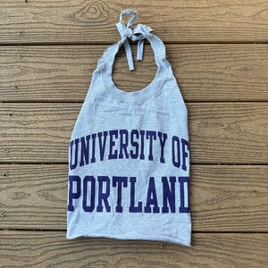 May include: A gray halter top made from a sweatshirt, featuring the text "UNIVERSITY OF PORTLAND" in navy blue block letters. The top is tied at the neck with a matching gray fabric.