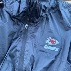 May include: A navy blue hooded windbreaker jacket with a full zipper closure. The jacket features the Kansas City Chiefs logo embroidered on the chest in red, white, and blue. The jacket is made of a lightweight, water-resistant material.