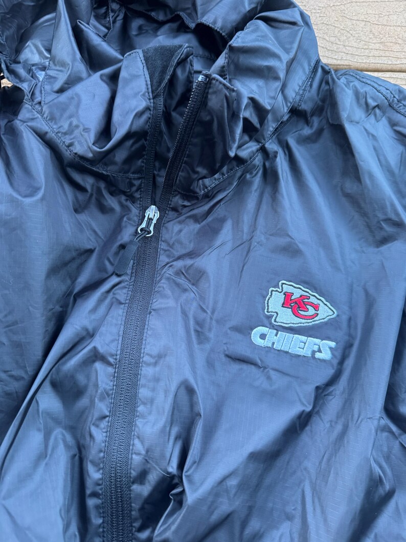 May include: A navy blue hooded windbreaker jacket with a full zipper closure. The jacket features the Kansas City Chiefs logo embroidered on the chest in red, white, and blue. The jacket is made of a lightweight, water-resistant material.