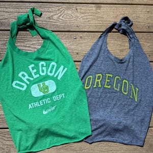 May include: Two sleeveless halter-style tops made from t-shirt material. The green top features the text OREGON, a UO logo, ATHLETIC DEPT., and a Nike swoosh. The grey heather top features the text OREGON in a lime green font. Both have tied necklines.