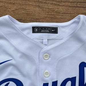 May include: A white baseball jersey with a textured fabric, featuring two white buttons at the collar. The interior neck label displays the MLB Genuine Merchandise logo, size M 10/12, Made in Vietnam, and a Nike swoosh logo. Blue lettering is visible.