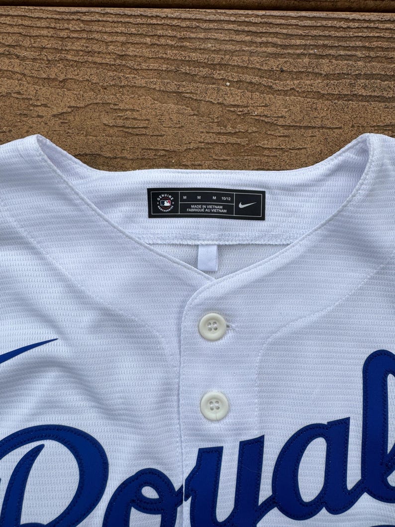May include: A white baseball jersey with a textured fabric, featuring two white buttons at the collar. The interior neck label displays the MLB Genuine Merchandise logo, size M 10/12, Made in Vietnam, and a Nike swoosh logo. Blue lettering is visible.