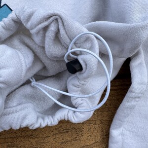 May include: A white sweatshirt featuring a drawstring hem with a black plastic toggle. The fabric has a soft, fleece-like interior texture. A portion of a graphic design with light blue shapes and the letters TM is visible at the top edge.