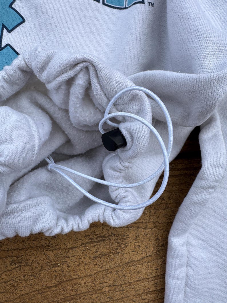 May include: A white sweatshirt featuring a drawstring hem with a black plastic toggle. The fabric has a soft, fleece-like interior texture. A portion of a graphic design with light blue shapes and the letters TM is visible at the top edge.