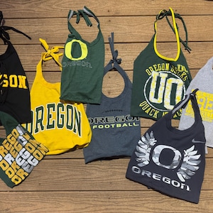 May include: Assorted halter tops in various colors, featuring the word "OREGON" and other university-related graphics. The tops are made from repurposed t-shirts and have tie closures.