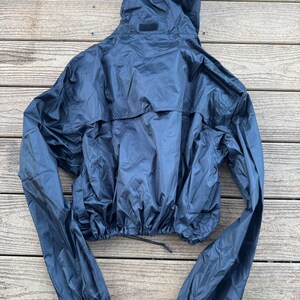 May include: A dark navy blue hooded jacket with long sleeves. The jacket has a drawstring at the waist and elastic cuffs. The hood is attached and has a small patch on the back. The jacket is made of a shiny, lightweight material.