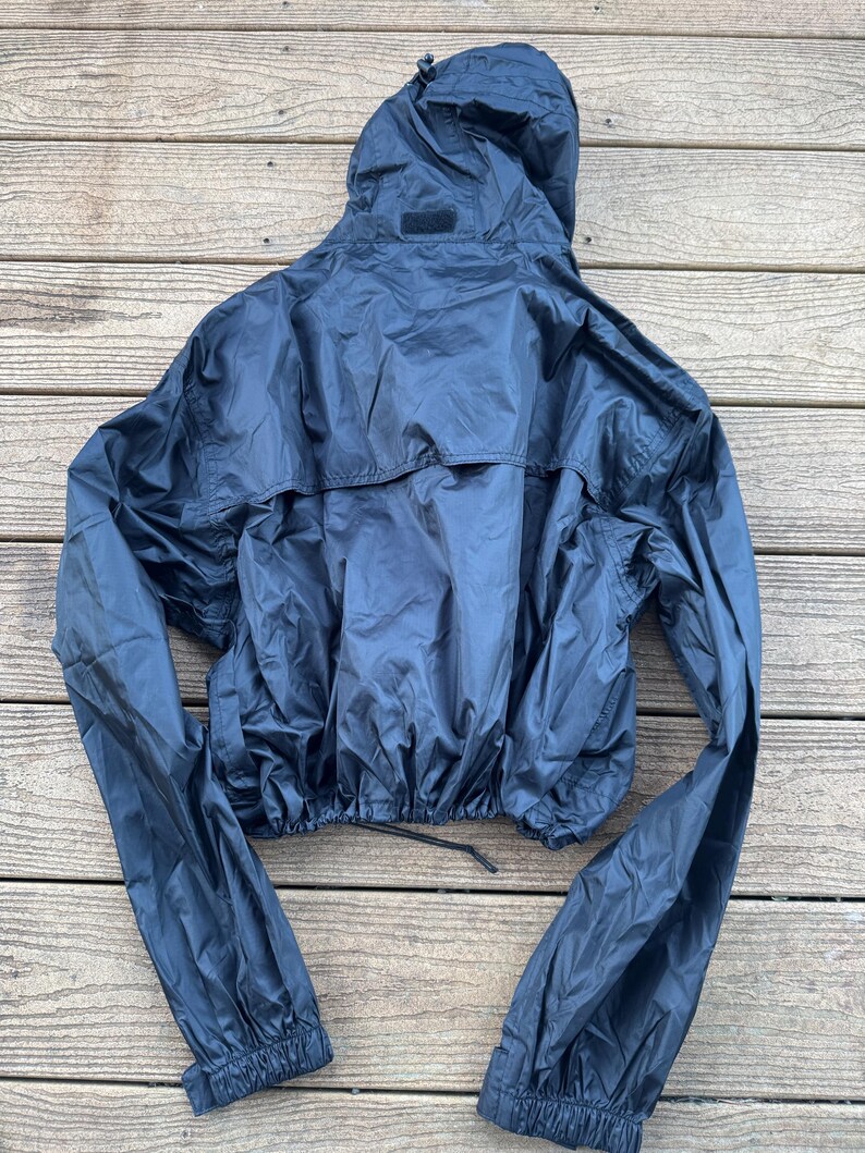 May include: A dark navy blue hooded jacket with long sleeves. The jacket has a drawstring at the waist and elastic cuffs. The hood is attached and has a small patch on the back. The jacket is made of a shiny, lightweight material.