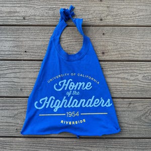 May include: A royal blue bib-style shirt with the text "UNIVERSITY OF CALIFORNIA Home of the Highlanders 1954 RIVERSIDE" in white and yellow. The shirt has a knotted tie at the top.