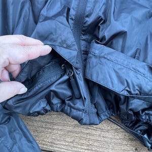 May include: A black nylon jacket with a zipper closure and a drawstring hem. The jacket has a pocket with a zipper. The jacket is on a wooden surface.
