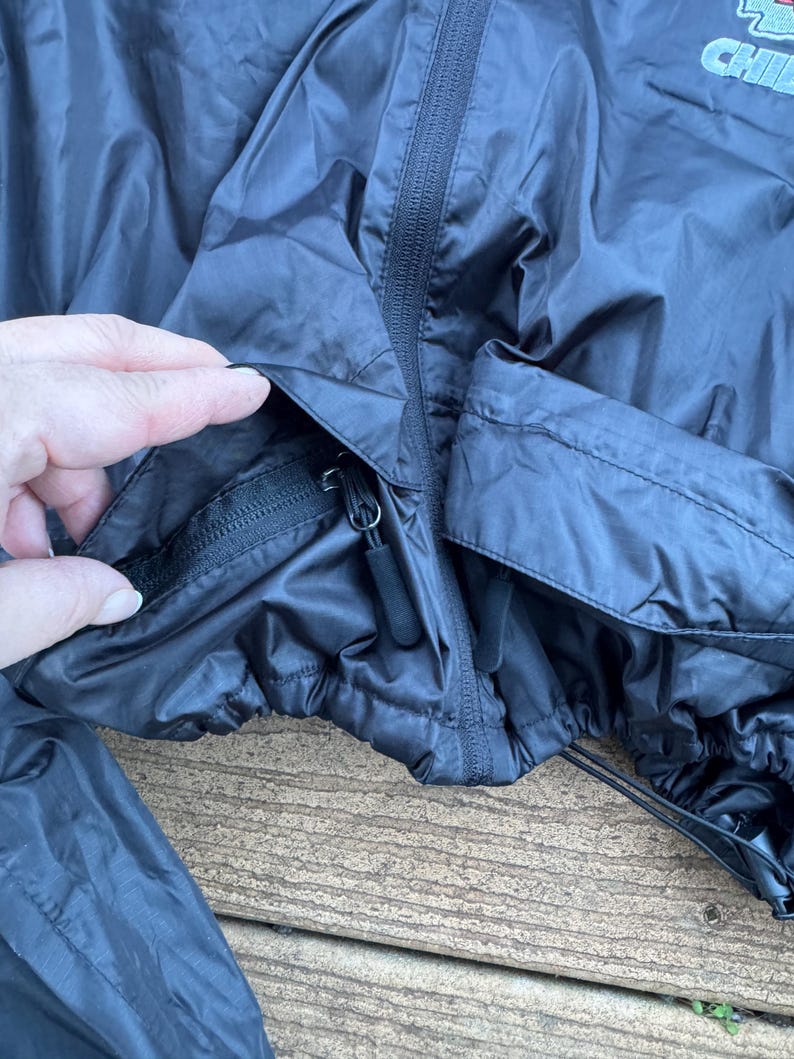 May include: A black nylon jacket with a zipper closure and a drawstring hem. The jacket has a pocket with a zipper. The jacket is on a wooden surface.