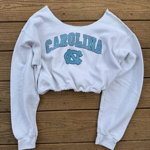 May include: A white long-sleeve cropped sweatshirt featuring a wide scoop neckline and an elasticized hem. The front displays the word CAROLINA in light blue block letters above a light blue interlocking NC logo, all printed in a collegiate style.
