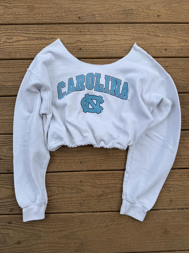 May include: A white long-sleeve cropped sweatshirt featuring a wide scoop neckline and an elasticized hem. The front displays the word CAROLINA in light blue block letters above a light blue interlocking NC logo, all printed in a collegiate style.