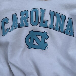 May include: A white sweatshirt featuring the word CAROLINA in large, light blue letters with a dark blue outline, positioned above a light blue interlocking NC logo. The design is printed on the front of the garment, which has a crew neckline.