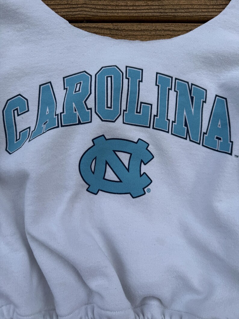 May include: A white sweatshirt featuring the word CAROLINA in large, light blue letters with a dark blue outline, positioned above a light blue interlocking NC logo. The design is printed on the front of the garment, which has a crew neckline.
