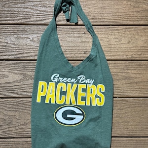 May include: A heather green halter top featuring a V-neckline and a tie closure at the neck. The front displays the text Green Bay PACKERS in yellow and white lettering, positioned above the official yellow and white G logo on a dark green background.